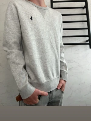 Grå sweatshirt från Polo Ralph Lauren - Klassisk ljusgrå sweatshirt från Polo Ralph Lauren med rund halsringning och diskret broderad logga på bröstet. Tröjan har ribbade muddar vid ärmslut och nederkant, perfekt för en avslappnad och stilren look.