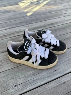 Svarta Adidas campus  - Säljer ett par klassiska svarta Adidas sneakers med vita ränder och breda vita skosnören. Skorna har använts med har tagits hand om och har mer kvar att ge, går säkert att fräscha upp ännu mer. Skokartong ingår🥰