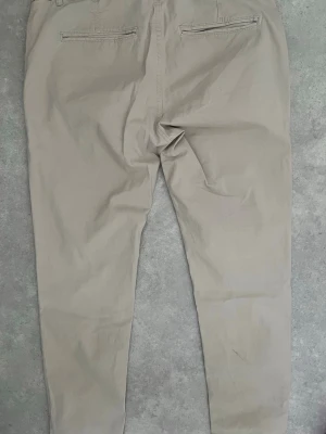 Beige chinos med smal passform - Snygga beige chinos med smal passform och två bakfickor med dragkedja. Tillverkade i ett mjukt bomullsmaterial som ger en clean och stilren look. Perfekta för dig som gillar en enkel men modern stil.