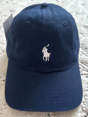 Mörkblå keps från Polo Ralph Lauren - Snygg mörkblå keps från Polo Ralph Lauren med klassisk vit broderad logga framtill och 'POLO' broderat bak. Justerbart spänne i metall baktill och böjd skärm. Tillverkad i bomull för en bekväm och stilren look.