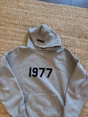Essentials fear of god 1977 hoodie - Säljer en grå hoodie från essentials med svart tryck '1977' på bröstet. Hoodien har en klassisk huva och en stor magficka framtill. Perfekt för en avslappnad och cool stil. Materialet är mjukt och bekvämt, troligen bomullsblandning.