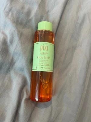 Pixi Glow Tonic 250ml ansiktstoner - Pixi Glow Tonic är en exfolierande ansiktstoner med 5% glykolsyra. Kommer i en cylinderformad plastflaska med mintgrönt lock och etikett. Innehållet är orangefärgat och flaskan rymmer 250 ml. Perfekt för dig som vill ha glow och fräsch hy.