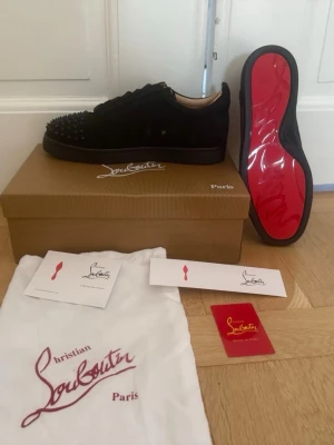 Svarta Louboutin sneakers med nitar - Säljer ett par svarta sneakers från Christian Louboutin med mattsvarta nitar på tån och den ikoniska röda sulan. Skorna är låga och har en lyxig känsla med mockaliknande material. Perfekta för dig som vill sticka ut med exklusiv design.