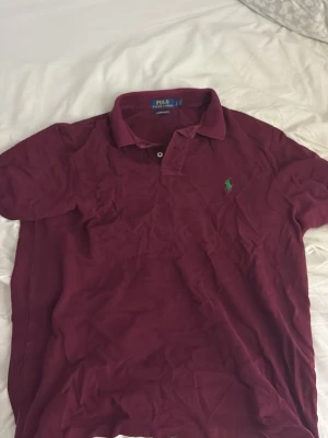 Vinröd pikétröja från Ralph Lauren - Säljer en vinröd pikétröja från Ralph Lauren med klassisk krage och två knappar vid halsen. Tröjan har en broderad grön logga på bröstet och är i modellen custom slim fit. Perfekt för en stilren och avslappnad look.