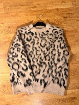 Beige leopardmönstrad stickad tröja - Säljer en beige stickad tröja med svart leopardmönster. Tröjan har rund halsringning, långa ärmar och ribbade muddar vid ärmslut och nederkant. Materialet ser mjukt och fluffigt ut, perfekt för vintern.                                         Aldrig använd(: