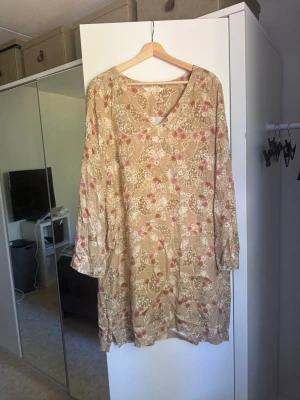 Beige blommig långärmad klänning - Söt guld klänning med blommigt mönster i vitt och rosa. Klänningen har v-ringning och långa ärmar, samt en lös och bekväm passform. Perfekt för sommarens varma dagar eller som en bohemisk touch till din garderob. Från märket Odd Molly i storlek 3 som enligt deras storlekguide motsvarar en storlek L. Mycket fint skick, jag hittar inga synliga anmärkningar eller defekter.  