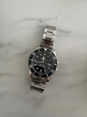 Klassisk silverfärgad klocka - Stilren klocka i silverfärgat metallarmband och svart urtavla med vita detaljer. Klockan har en rund form och tydliga markeringar på boetten. Perfekt accessoar för dig som gillar en tidlös look.