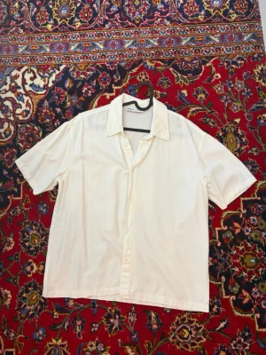 Vit kortärmad skjorta från Bershka - Säljer en stilren vit skjorta från Bershka med korta ärmar och klassisk krage. Skjortan har knappar framtill och är tillverkad i ett lätt och luftigt material, perfekt för varmare dagar. Enkel att matcha med olika outfits.
