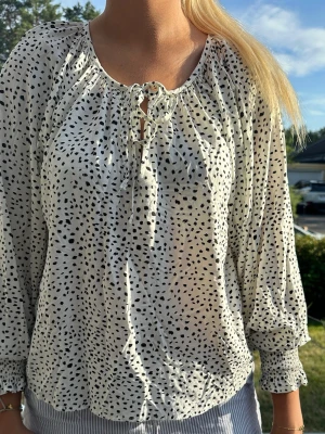 Prickig blus - Säljer en luftig vit blus med svarta prickar och knytning vid halsen. Blusen har långa, ballongformade ärmar med resår vid ärmsluten. Köpt på Lindex i storlek S