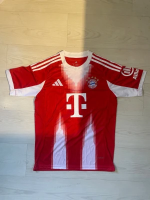 FC Bayern München matchtröja röd - Snygg röd FC Bayern München matchtröja från Adidas med vita detaljer och klubbmärke på bröstet. Stort T-sponsormärke framtill och Musiala 10 tryckt på ryggen. Tillverkad i lätt och ventilerande material, perfekt för fotbollsträning eller match. Säljer billigt för den passade inte