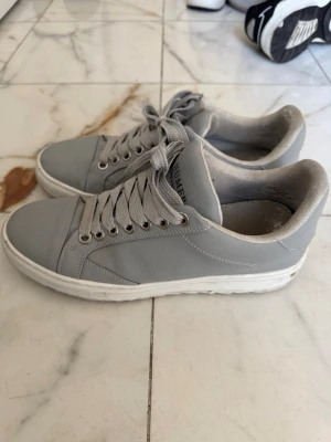 Numeris - Snygga grå sneakers från Numeris med vita sulor och matchande grå skosnören. Skorna har en stilren design i slätt material och lågt skaft. Perfekta för dig som gillar en clean och enkel look till vardagsoutfiten.