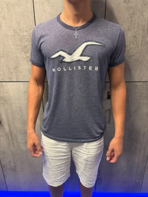 *UNIK* Blå Hollister t-shirt - Säljer en blå t-shirt från Hollister med stor vit logga och text på bröstet. T-shirten har rund halsringning och korta ärmar med mörkare kant. Perfekt för en avslappnad och stilren look. Perfekt om du vill se lite arkiv ut😂