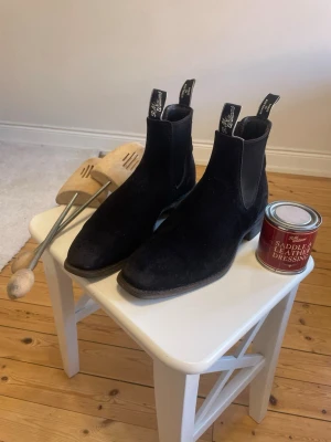 RM Williams | Svart mocka, UK 9,5 G | Made in Australia -  Klassiska chelseaboots från RM Williams i svart mocka, Made in Australia. UK 9,5 G (ca EU 43,5–44).  Normalt bruksslitage, mockan har blankare partier (se bild). Elastiken hel med god spänst. Gummisulorna har gott om mönster kvar. Alltid förvarade med skoblock. Sula och läderkanter vårdade med RM Williams Saddle & Leather Dressing.  Ingår: läderfettet (200 g, delvis använt), skoblock och ett extra par innersulor.  Avhämtning Stockholm eller Göteborg, kan skickas. Hör av dig vid frågor.