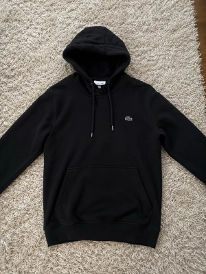 Svart hoodie från Lacoste - Snygg svart hoodie från Lacoste med klassisk krokodillogo på bröstet. Hoodien har huva med dragsko och en stor magficka framtill. Tillverkad i mjukt material och har ribbade muddar vid ärmslut och nederkant. Perfekt för en chill och stilren look.