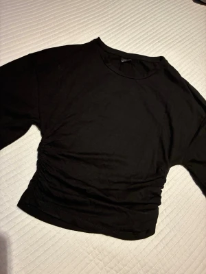 Svart croppad topp med rynkade sidor storlek S - En svart croppad topp med rund halsringning och rynkade detaljer på sidorna. Tillverkad i mjukt och stretchigt material, perfekt för en avslappnad och trendig look. Passar till både jeans och kjol. Storlek S från Gina.