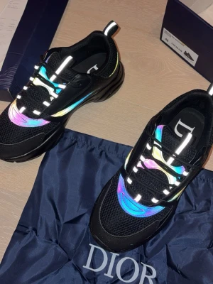 Dior sneakers med reflexdetaljer - Säljer ett par svarta Dior sneakers med färgglada reflexdetaljer i blått, rosa och gult. Skorna har meshpaneler, vita reflekterande snören och en chunky sula. Perfekta för dig som vill sticka ut med en exklusiv och modern look.