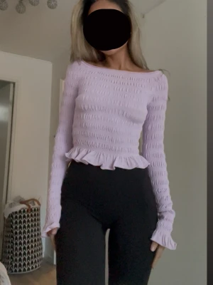 Ljuslila smockad topp med volang - Supersöt ljuslila topp med smockad struktur och volangkant nedtill. Långärmad modell med figurnära passform och stretchigt material.