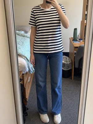 Navyvit randig t-shirt - Klassisk t-shirt med navy och vita horisontella ränder. Modellen har rund halsringning och korta ärmar. Perfekt för en avslappnad och stilren look. Materialet ser ut att vara mjukt och bekvämt, troligtvis bomull.