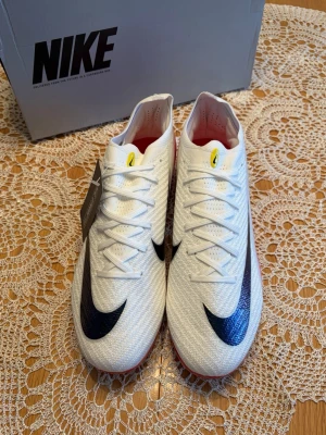 Vita Nike Mercurial fotbollsskor - Snygga vita Nike Mercurial fotbollsskor med svart Swoosh på ovansidan och röda detaljer på sulan. Skorna har en modern, strömlinjeformad design och är tillverkade i ett lätt, syntetiskt material för bästa bollkontroll och snabbhet på planen.