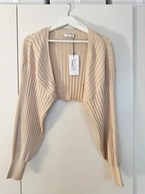 Beige ribbad bolero från Stockh lm - Snygg beige ribbad bolero från Stockh lm med öppen front och långa ärmar. Tillverkad i mjukt stickat material som ger en avslappnad men trendig look. Perfekt att slänga över en topp för extra stilpoäng.