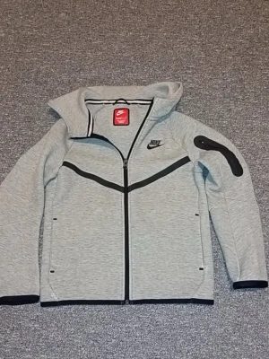 Grå Nike hoodie med dragkedja - Snygg grå hoodie från Nike med hel dragkedja framtill och svarta detaljer. Har två sidofickor med dragkedja och en extra ficka på ärmen. Nike-logga på bröstet och sportig design. Perfekt för en avslappnad streetwear-look.
