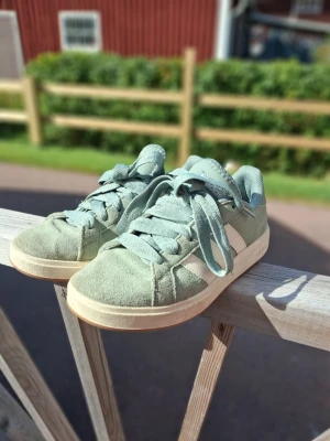 Gröna Adidas sneakers i mocka - Snygga gröna Adidas sneakers i mocka med vita detaljer och klassiska ränder på sidan. Skorna har breda gröna skosnören och vit sula. Perfekta för dig som vill ha en stilren och trendig look till vardags.