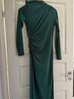 Grön långklänning med rynkade detaljer - Snygg lång grön klänning med långa ärmar och rynkade detaljer längs sidan. Klänningen har en elegant och figurnära passform, perfekt för dig som vill sticka ut. Tillverkad i ett mjukt och följsamt material.