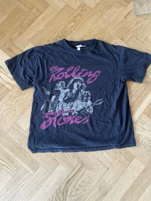 Svart Rolling Stones t-shirt från H&M - Cool svart t-shirt från H&M med Rolling Stones-tryck i rosa och vitt framtill. Klassisk rund hals och korta ärmar. Perfekt för dig som gillar bandtröjor och vill ha en avslappnad stil.
