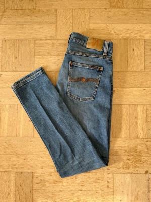 Blå Nudie jeans i modell Lean Dean (slim fit) - Säljer ett par klassiska blå jeans från Nudie Jeans med snygga slitningar framtill. Jeansen har fem fickor, orange sömmar och Nudie-logga på bakfickan. Perfekta för en avslappnad och trendig look. W30 L32 Hojat bara till vid frågor!😁🙌