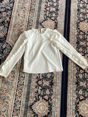 Vit blus med volangdetaljer - Söt vit blus med långa ärmar och volangdetaljer längs axlar och ärmar. Tillverkad i ett strukturerat, mjukt tyg som ger en lyxig känsla. Rund halsringning och knapp i nacken för enkel påklädning. Perfekt för att lyfta vilken outfit som helst.
