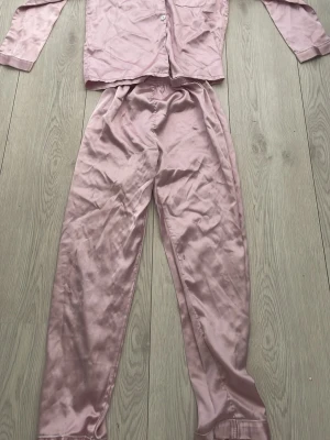 Ljusrosa satinpyjamas set - Säljer ett ljusrosa pyjamas set i satin-look med långärmad skjorta och långa byxor. Materialet är 97% polyester och 3% elastan vilket gör det mjukt och stretchigt. Skjortan har knappar framtill och byxorna har resår i midjan för extra komfort.