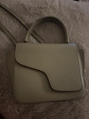 Grön ATP väska - Stilren beige handväska i slätt konstläder med ett unikt asymmetriskt lock. Väskan har både handtag och axelrem för olika bärstilar. Perfekt för dig som gillar minimalistisk design och vill ha en väska som sticker ut lite extra.