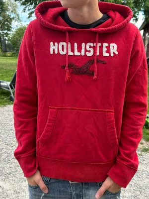 Röd hoodie från Hollister - Snygg röd hoodie från Hollister med stor magficka och broderad logga samt text på bröstet. Hoodien har dragsko i huvan och ribbade muddar vid ärmar och nederkant. Perfekt för en avslappnad och trendig stil. Den är lite sliten