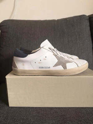 Golden goose superstars - Snygga vita sneakers med beige stjärna på sidan och GGDB/SSSTAR-tryck. Skorna har beige sula och mörkblå detalj vid hälen. Klassisk låg modell med vita snören och perforerade detaljer på tungan. Tillverkade i läder och mocka för en trendig look.