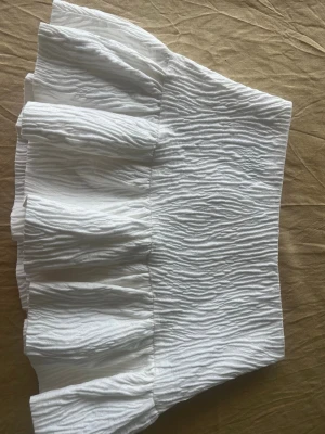 Vit kortkjol med volang - Supersöt vit kortkjol med volangkant och strukturerat tyg som ger en trendig look. Kjolen har en hög midja och är perfekt för dig som vill ha en fräsch och somrig stil. Har inbyggda shorts.  Det är storlek L men passar mig som i vanliga fall har M och S. Pris kan diskuteras! 