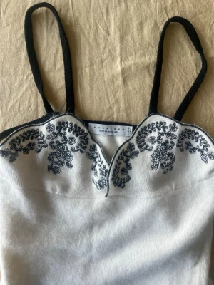 Vit linnetopp med broderi - Supersnygg vit linnetopp med svarta axelband och svart broderad detalj upptill. Toppen har en unik skuren kant vid bysten och tunna axelband. Perfekt att styla med jeans eller kjol för en trendig look. Pris kan diskuteras! 