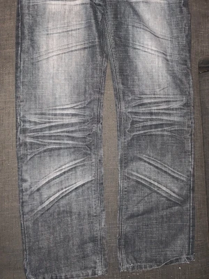 Grå slitna jeans med raka ben - Säljer ett par grå jeans med slitningar och markerade tvättade detaljer över låren och knäna. Jeansen har raka ben och en avslappnad passform. Materialet är klassisk denim och färgen är en cool gråton med ljusare partier.