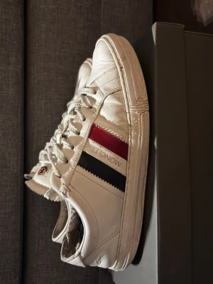 Vita Moncler sneakers med röd/blå detalj - Snygga vita sneakers från Moncler med klassiska röd och blå ränder på sidan och logga på plösen. Skorna har vita snören och en låg profil. Tillverkade i läder med gummisula. Perfekta för dig som vill ha en clean och stilren look. Dom har ett litet hål på sidan där av priset 