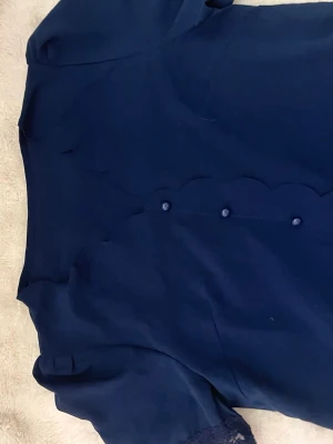 Mörkblå blus med vågig kant - Snygg mörkblå blus med korta ärmar och dekorativa vågiga kanter längs knappraden. Blusen har klädda knappar framtill och är gjord i ett mjukt, tunt tyg som ger en elegant känsla. Perfekt att styla med jeans eller kjol för en trendig look.
