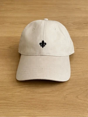 Morris keps  - Snygg beige keps med en liten svart broderad detalj framtill. Klassisk böjd skärm och justerbart spänne bak för perfekt passform. Tillverkad i bomullstyg som andas och passar till de flesta outfits.