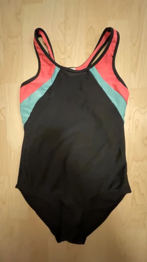 Svart baddräkt från KappAhl - Snygg svart baddräkt från KappAhl med sportig design. Har kontrastpaneler i turkos och korallrött vid axlarna och sidorna. Klassisk modell med dubbla axelband och rundad halsringning. Perfekt för simträning eller strandhäng.