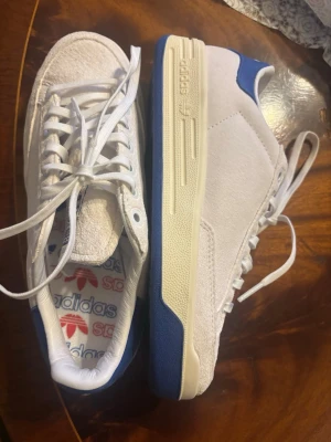 Rod laver Vita och blå Adidas sneakers - Rod laver skor, aldrig använt. Snygga vita sneakers från Adidas med blå detaljer på häl och sula. Skorna har en klassisk låg siluett, vita skosnören och ovandel i mocka och textil. Adidas-logga syns på innersulan. Perfekta för en clean och sportig look.