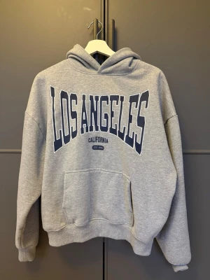 Grå hoodie med Los Angeles-tryck - Säljer en grå hoodie med stor text 'LOS ANGELES CALIFORNIA' i mörkblått framtill. Hoodien har huva, magficka och ribbade muddar vid ärmar och nederkant. Perfekt för en avslappnad och cool stil. Materialet känns mjukt och bekvämt, troligen bomullsblandning.