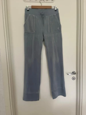 Blå velour mjukisbyxor Juicy Couture  - Säljer ett par ljusblå mjukisbyxor i velour med raka ben och breda muddar. Byxorna har stora framfickor, bakfickor med knapp och dragsko i midjan. Perfekta för chill dagar och riktigt sköna att ha på sig.
