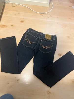 Mörkblå bootcut jeans med brodyr - Snygga mörkblå jeans med bootcut passform och dekorativa broderier på bakfickorna. Jeansen har kontrastsömmar och klassisk femficksdesign. Perfekta för dig som vill ha en unik detalj på dina jeans.