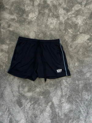 Svarta sportiga shorts med tryck - Snygga svarta shorts med vitt 1891-tryck på benet och diskreta ljusblå ränder längs sidorna. Shortsen har snörning i midjan och är perfekta för träning eller chill. Tillverkade i ett mjukt och lätt syntetmaterial som andas.