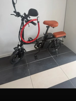 Svart elcykel med brunt säte - Säljer en svart elcykel med två bruna sadlar och robust ram. Cykeln har spegel, stötdämpare bak, batteri under sadeln och ett rött lås runt styret. Perfekt för smidig transport i stan och enkel att parkera tack vare stödet.