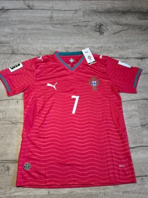 Cristiano Ronaldo Portugal nummer 7 - Snygg röd fotbollströja från Puma med Portugals landslagsemblem och nummer 7 på bröstet. Tröjan har vita detaljer, V-ringning med gröna inslag och vågiga linjer över hela tyget. Perfekt för match eller träning.