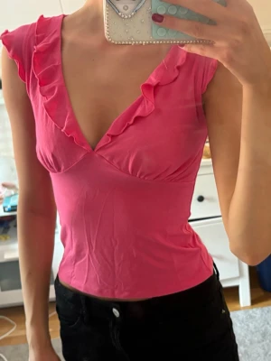 Rosa volangtopp från Gina Tricot - Säljer en söt rosa topp från Gina Tricot i storlek XS. Toppen har volangdetaljer längs den djupa v-ringningen och är ärmlös. Materialet är mjukt och stretchigt, perfekt för dig som gillar färg och vill sticka ut lite extra.