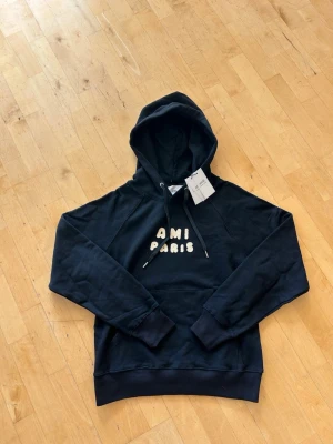 Svart hoodie från AMI Paris - Snygg svart hoodie från AMI Paris med vit broderad text på bröstet. Klassisk huva med dragsko och magficka framtill. Tillverkad i mjukt material som känns skönt mot huden. Perfekt för en avslappnad och trendig look.